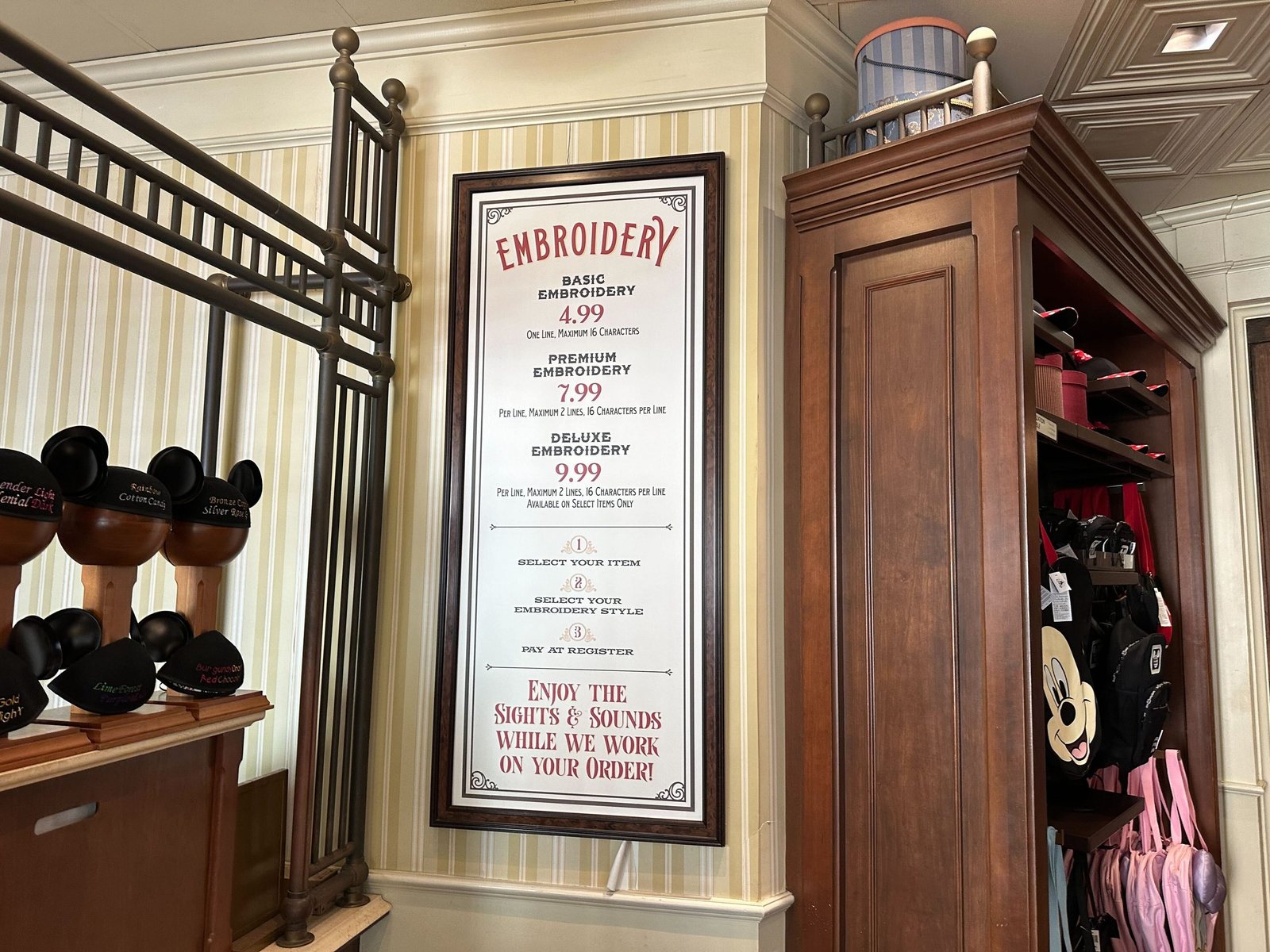 The same framed Embroidery sign at Curtain Call Collectibles in April 2026, listing Basic Embroidery 4.99, Premium Embroidery 7.99 and Deluxe Embroidery 9.99, beside a wooden cabinet holding Mickey backpacks, with sample Mickey ear hats on display stands at left