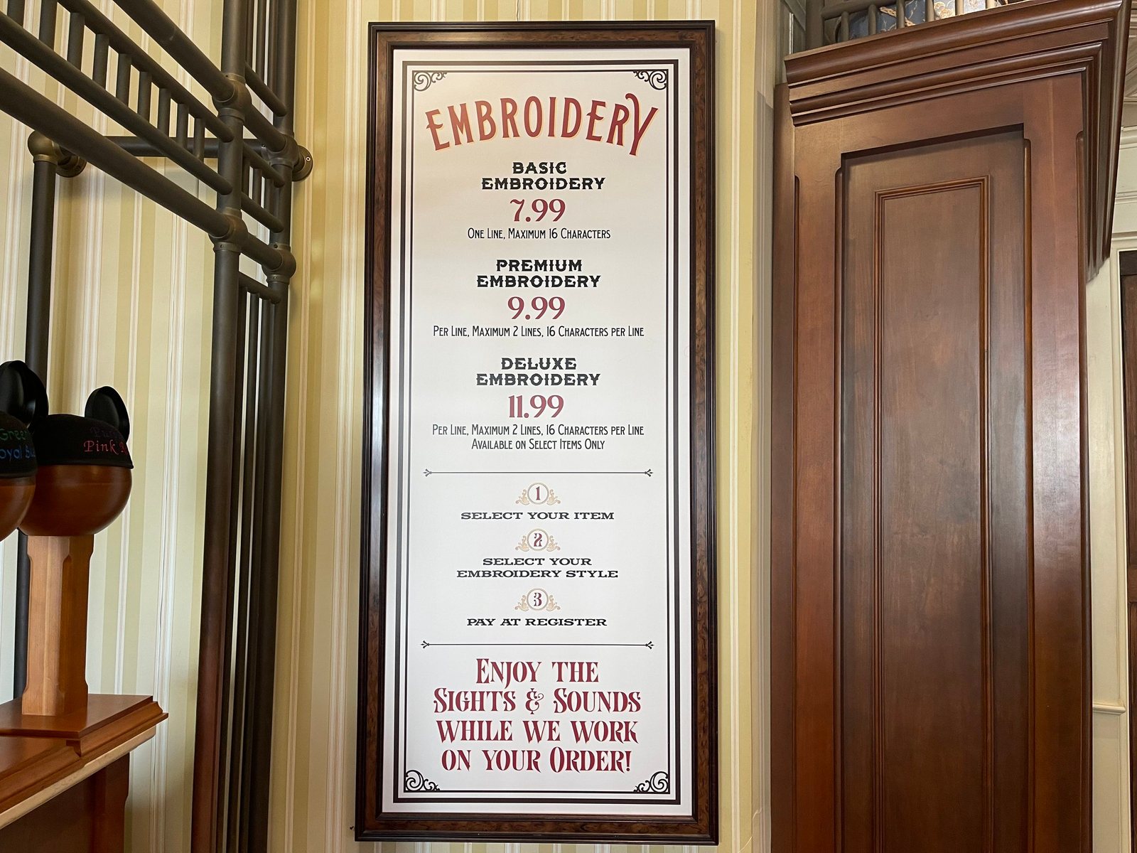 A framed Embroidery price sign on a striped wall at Curtain Call Collectibles, listing Basic Embroidery 7.99, one line, maximum 16 characters; Premium Embroidery 9.99 per line, maximum 2 lines; and Deluxe Embroidery 11.99 per line, available on select items only, with a Mickey ear hat display at left