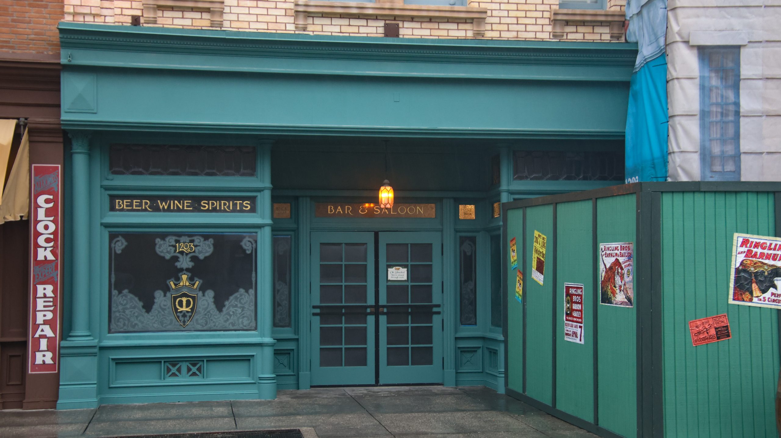 The freshly painted blue-green Finnegan's entrance, with Beer, Wine, Spirits and Bar and Saloon lettering in gold, an etched window with a crowned crest and the number 1203, a lit lantern over closed double doors, no Finnegan's sign or awning, and green construction walls with circus posters at right