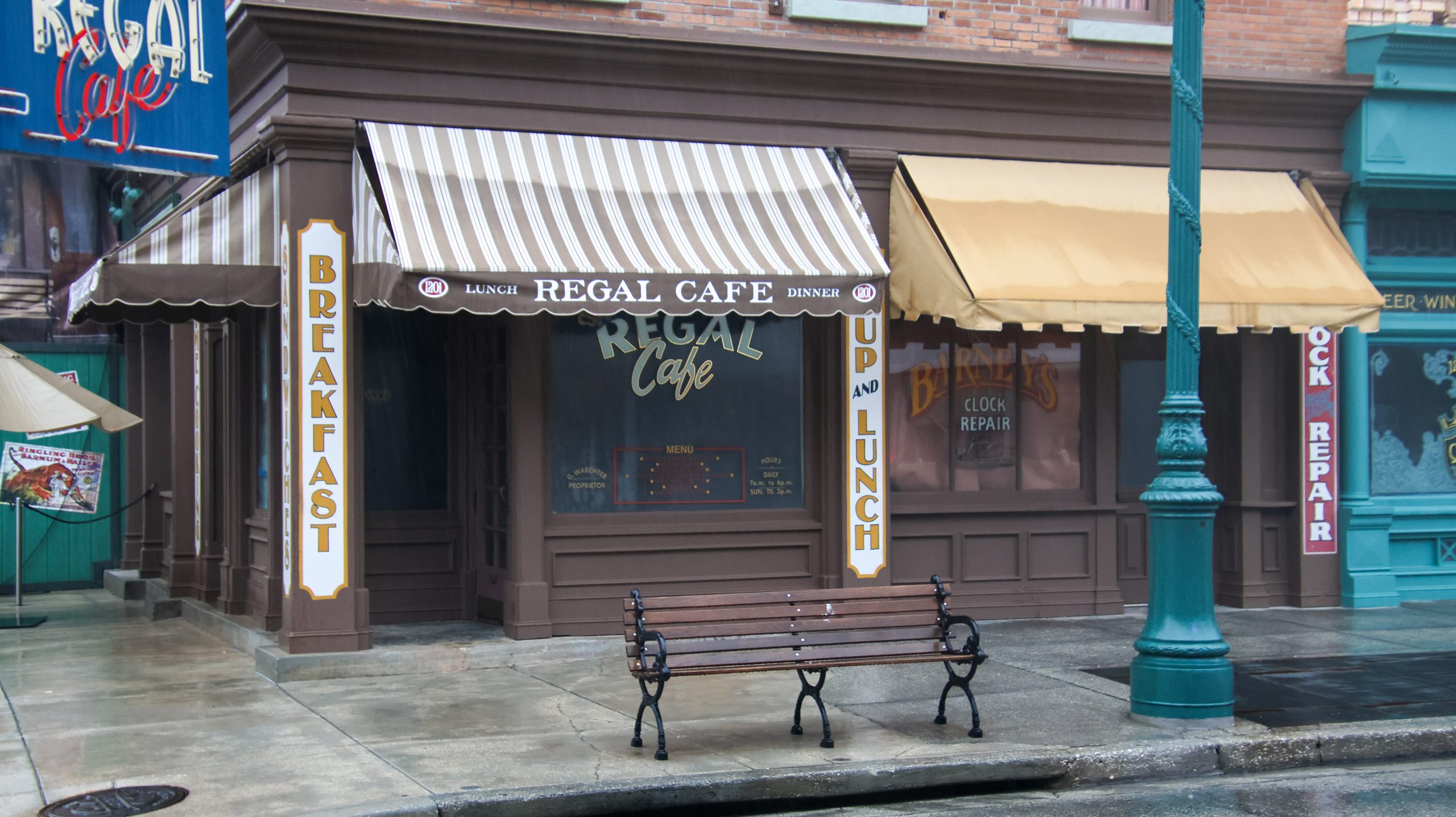 The Regal Cafe corner storefront in Universal Studios Florida's New York area, painted brown, with a beige and brown striped awning reading Lunch, Regal Cafe, Dinner, tall Breakfast and Soup and Lunch signs on the pillars, a solid tan awning over Barney's Clock Repair next door, and a park bench on the wet sidewalk