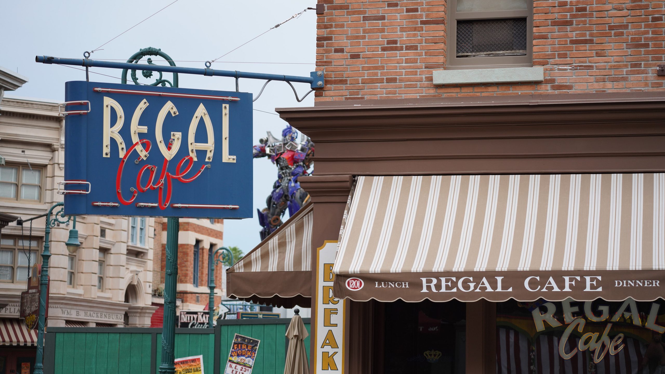 A blue hanging Regal Cafe sign with cream block letters and red script lettering, mounted on a bracket off the corner of a brick building, beside a beige and brown striped Regal Cafe awning, with green construction walls and an Optimus Prime statue in the background