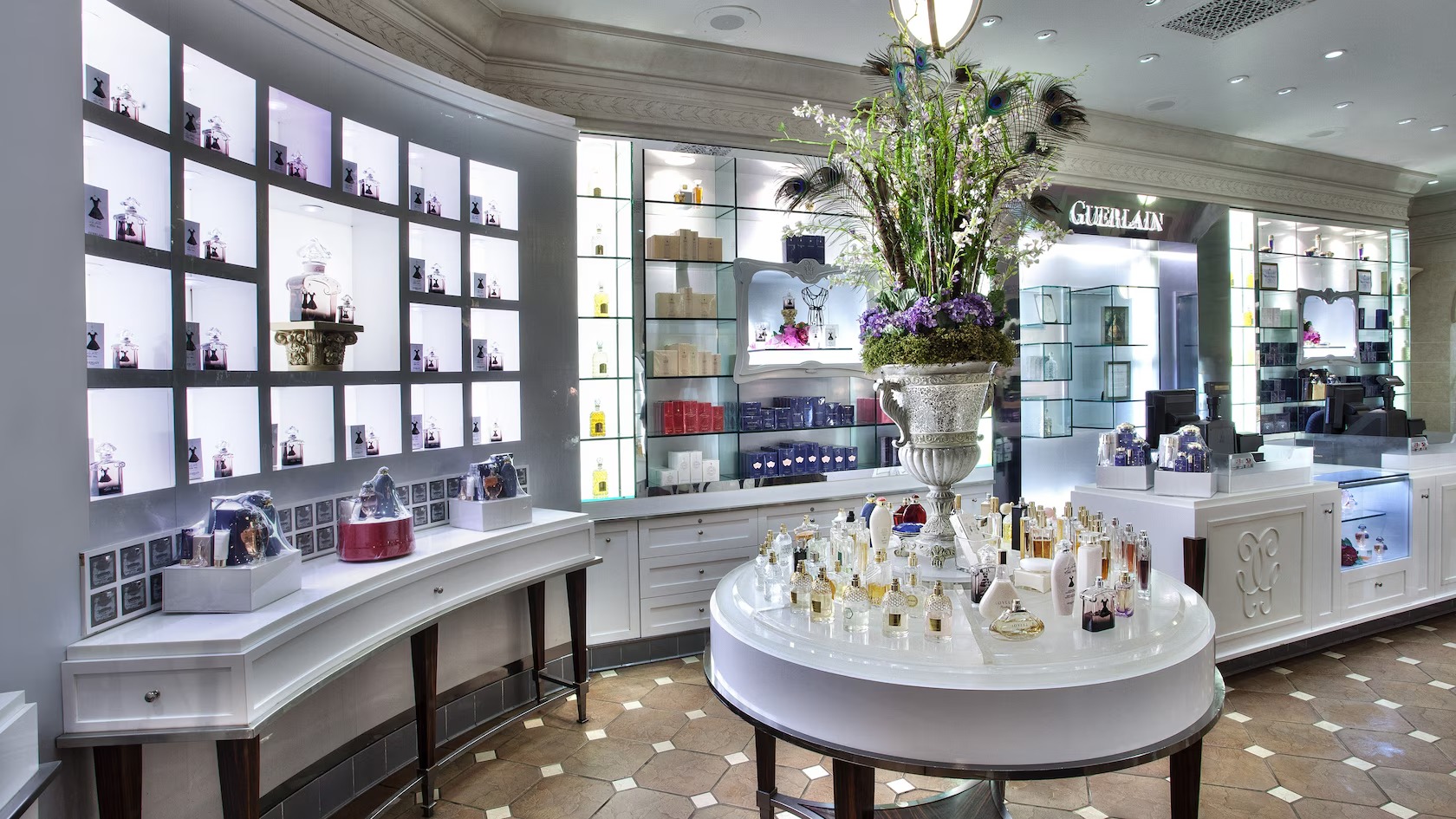 The interior of La Signature before its 2026 remodel, with backlit white cubby shelving holding perfume bottles, glass shelves along the back wall under a GUERLAIN sign, a round white display table of fragrance bottles beneath a tall floral arrangement with peacock feathers, and brown octagonal floor tiles