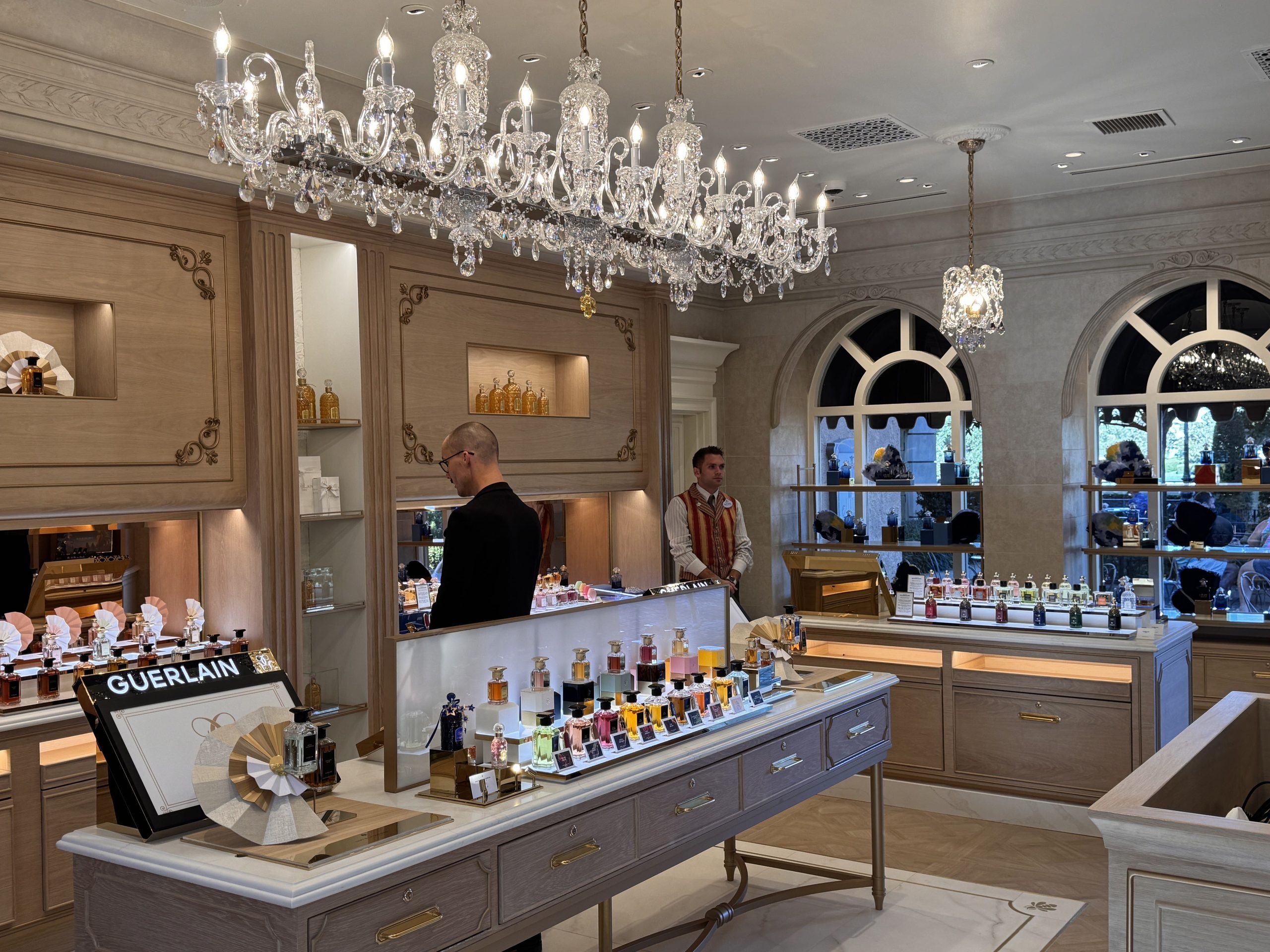 Inside the remodeled La Signature, a long crystal chandelier hangs over a white-topped display table of Guerlain fragrance bottles with a black GUERLAIN sign, light oak wall panels with gold scrollwork and lit niches of amber bottles behind, arched windows at right, and a cast member in a striped vest