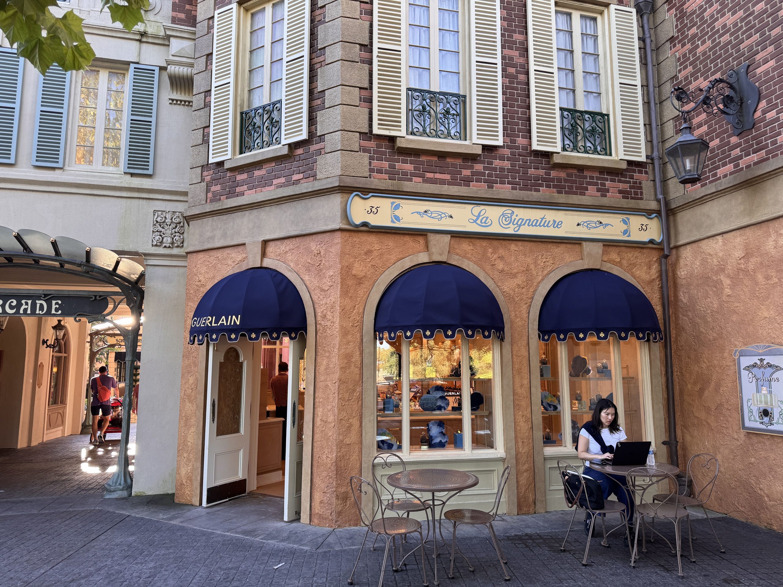 The corner La Signature storefront in EPCOT's France pavilion, with a cream La Signature 35 sign above three arched openings under navy blue awnings, one awning reading GUERLAIN over an open white door, and bistro tables and chairs on the pavement outside