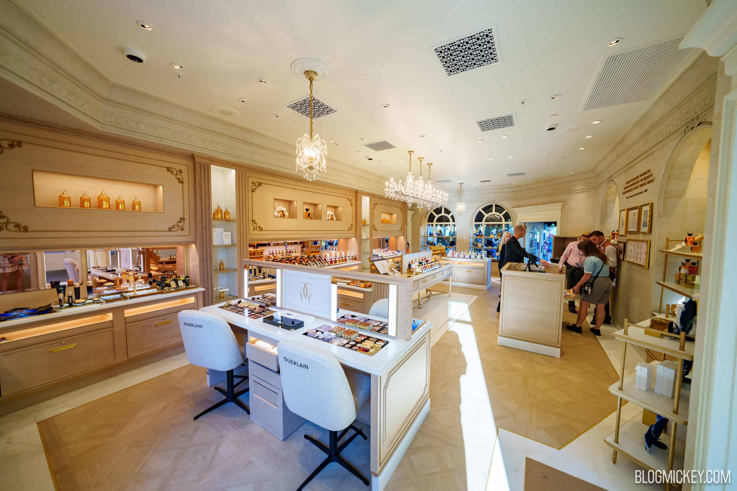 A wide view of the remodeled La Signature, with a white makeup vanity and two cream chairs embroidered GUERLAIN in the foreground, a parquet wood floor with white marble borders, several crystal chandeliers, oak wall cabinets lined with products, framed pictures on the right-hand wall and guests and cast members near the checkout