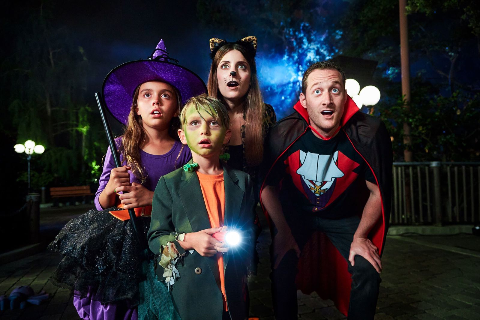 A family in Halloween costumes at night, a girl in a purple witch hat and dress, a boy with green face paint holding a flashlight, a woman with cat ears and whiskers and a man in a vampire cape, all staring wide-eyed at something out of frame