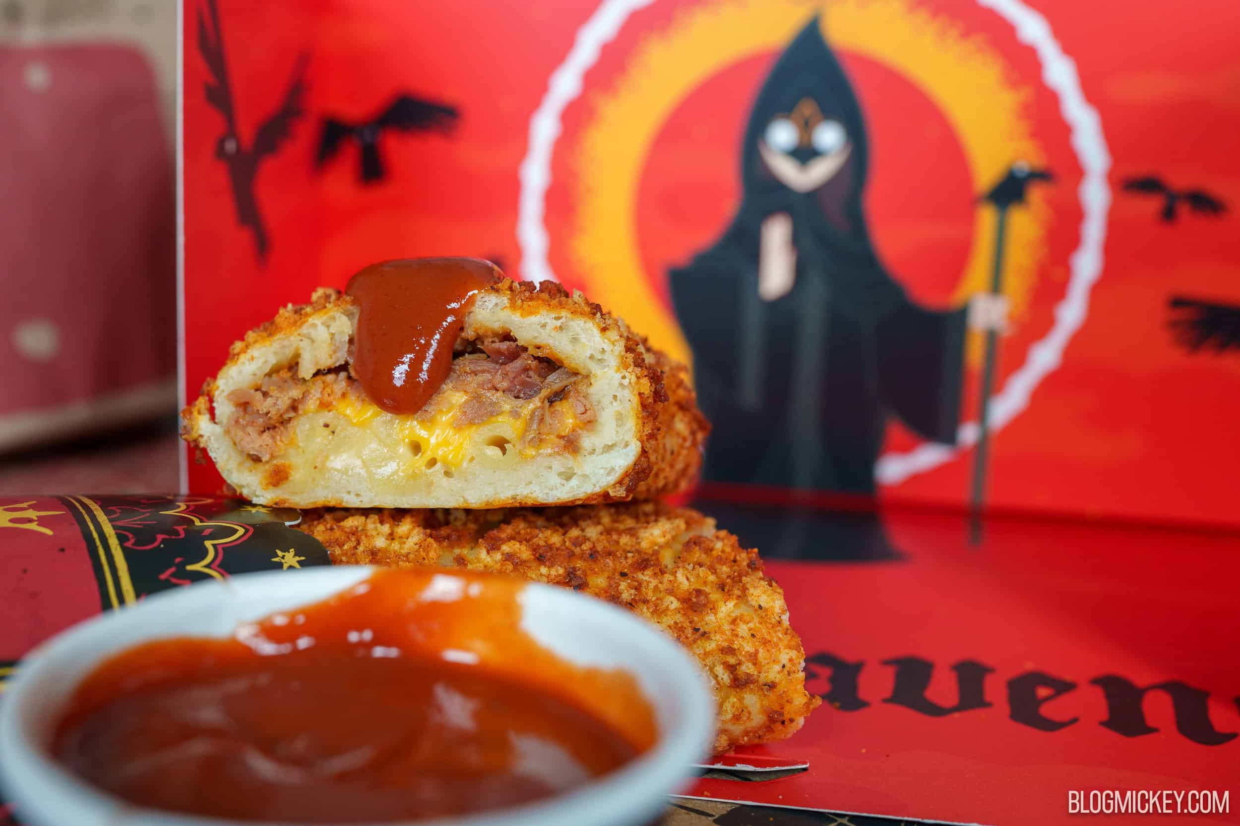 A golden, crumb-coated Coffyn cut in half to show shredded meat, macaroni and melted cheese inside, topped with a spoonful of red-brown dipping sauce, with a cup of the sauce in front and a red Six Ravens box printed with a hooded figure and flying ravens behind