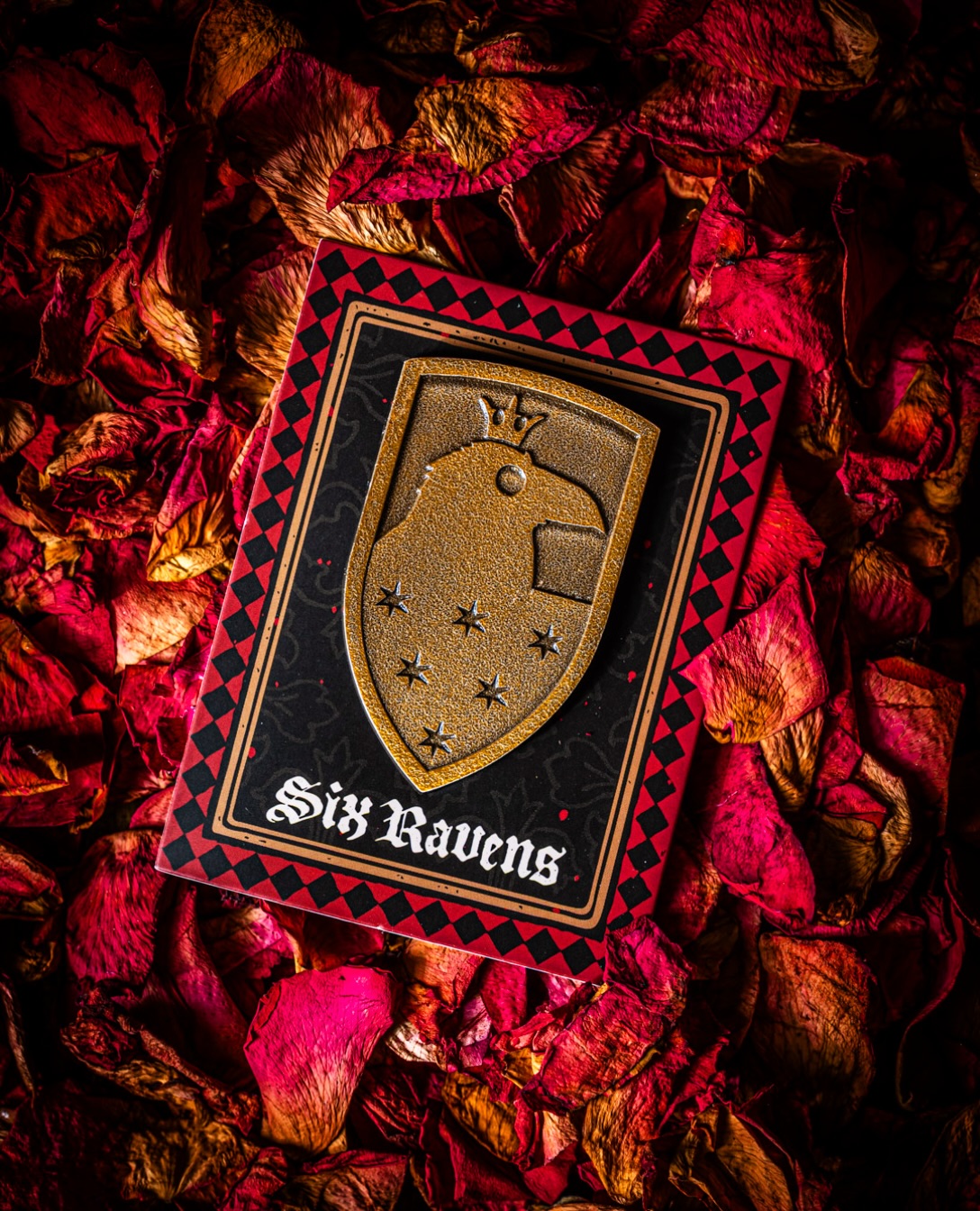 A gold shield-shaped pin showing a crowned raven's head above six stars, mounted on a black backing card with a red and black diamond-checked border and the words Six Ravens in white gothic lettering, photographed on a bed of red rose petals