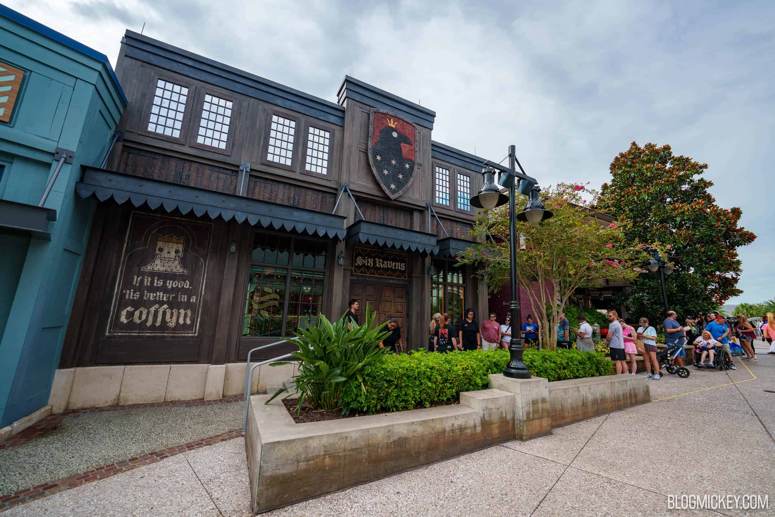 The dark timber Six Ravens storefront at Disney Springs, with a red shield showing a crowned black raven and six stars high on the facade, a Six Ravens sign over wooden double doors, a wall mural reading If it is good, 'tis better in a coffyn, and a line of guests along the planter outside