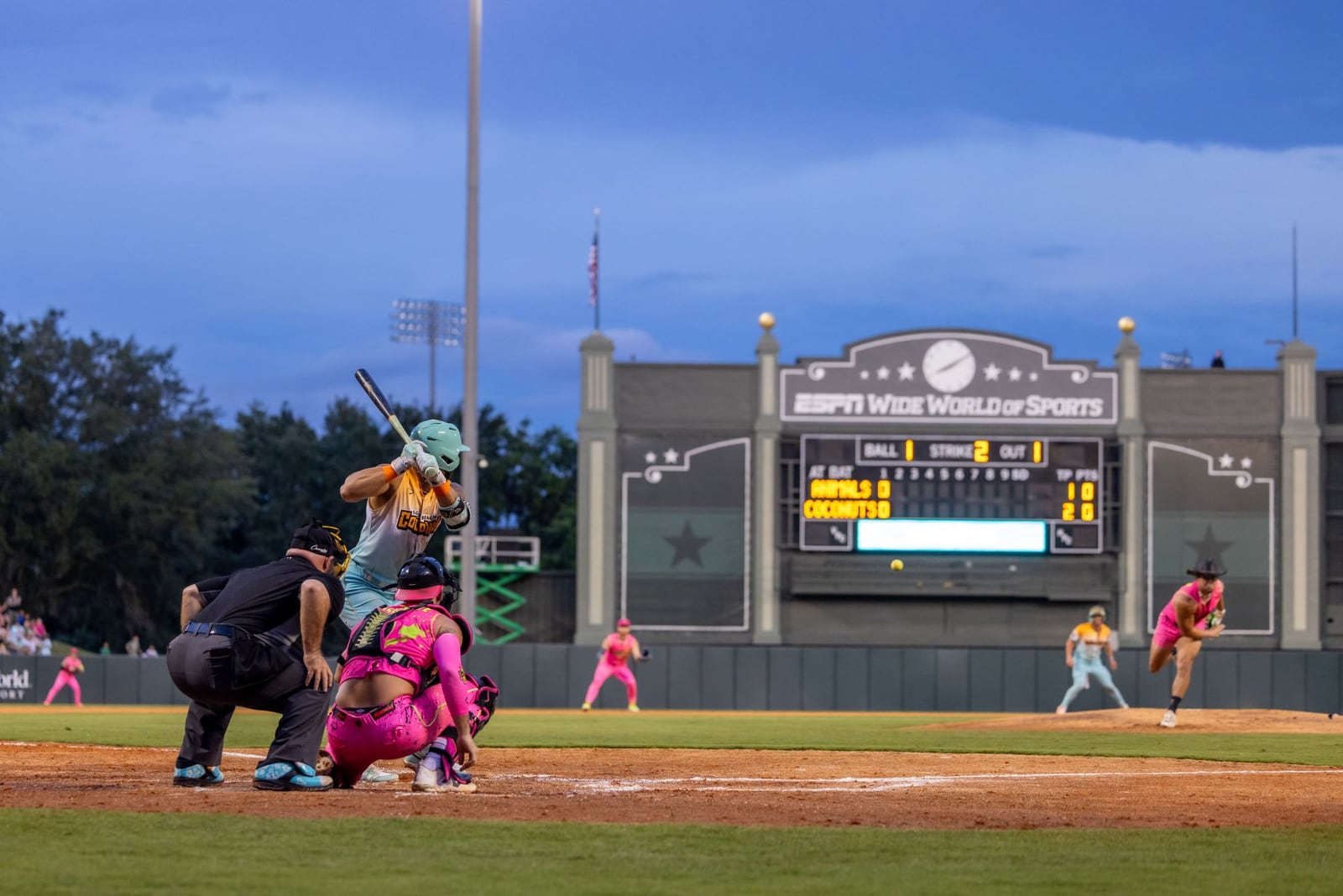A Loco Beach Coconuts batter in a mint helmet waits for a pitch while a Party Animals catcher in pink and an umpire crouch behind him at dusk, with the ESPN Wide World of Sports scoreboard in the outfield listing the Animals and Coconuts