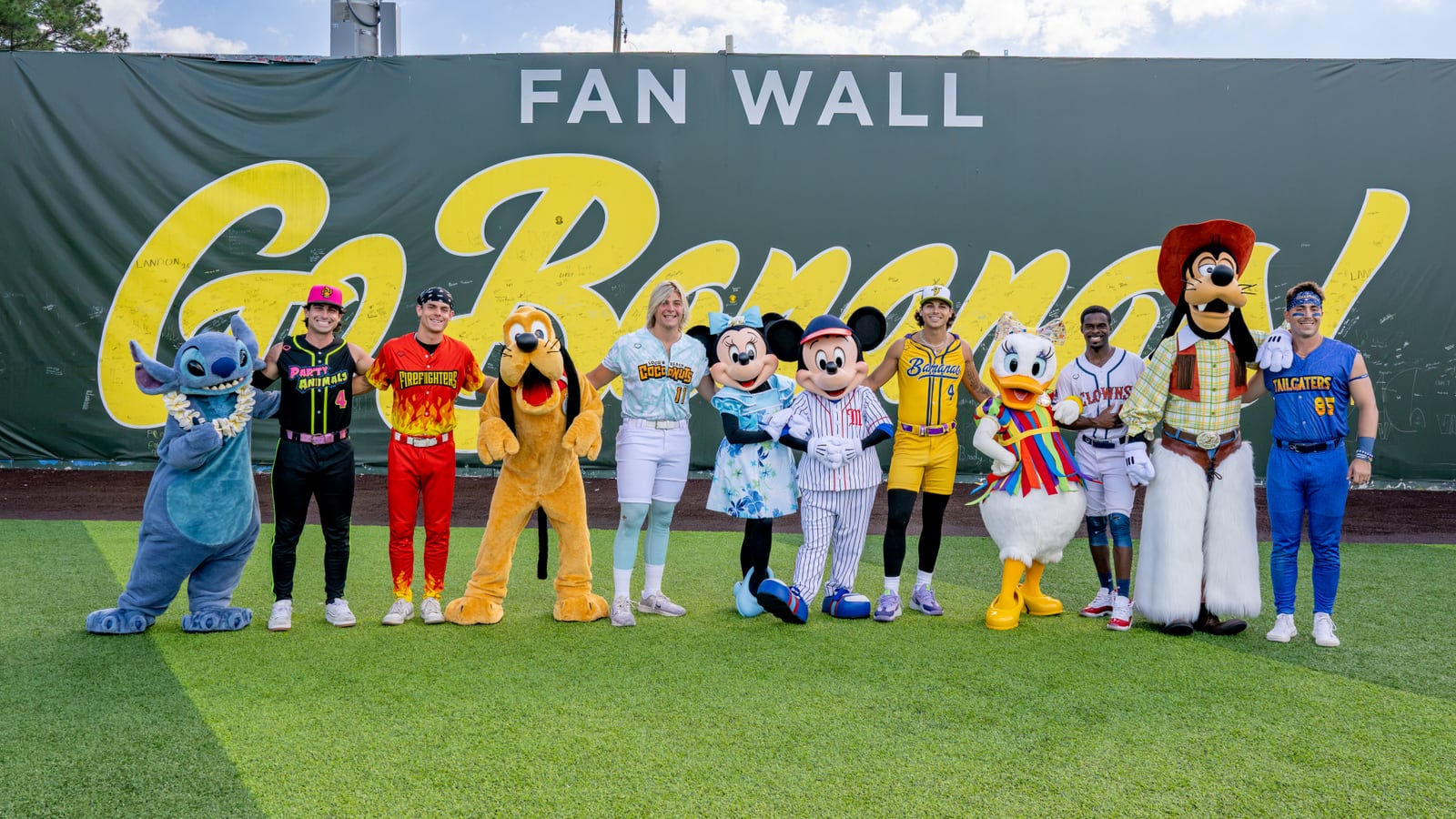 Stitch, Pluto, Minnie Mouse, Mickey Mouse in a pinstripe baseball uniform, Daisy Duck and Goofy in cowboy gear line up on artificial turf with six Banana Ball players in Party Animals, Firefighters, Loco Beach Coconuts, Savannah Bananas, Indianapolis Clowns and Texas Tailgaters uniforms, in front of a green Fan Wall reading Go Bananas!