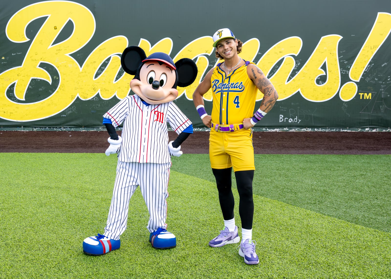 Mickey Mouse in a white pinstripe baseball uniform and cap stands hands on hips beside a Savannah Bananas player in a yellow number 4 jersey and shorts, in front of a green wall with giant yellow Bananas! lettering covered in signatures