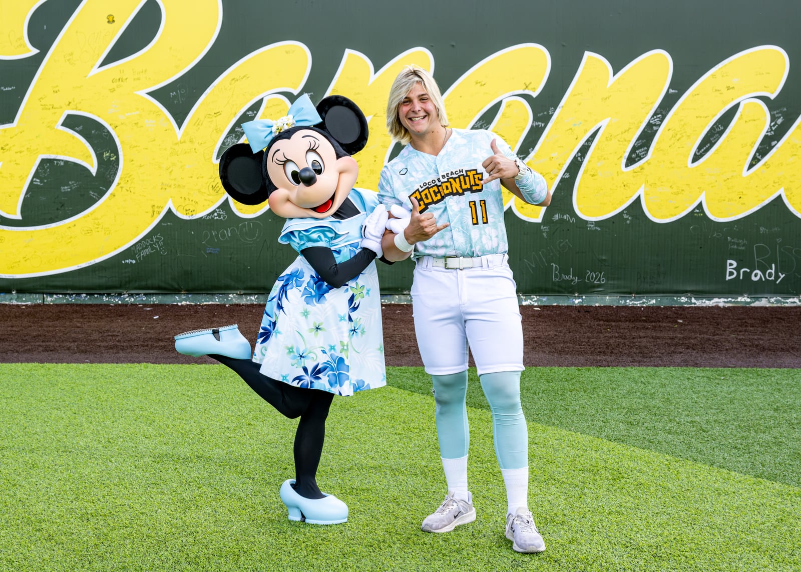 Minnie Mouse in a blue floral dress kicks up one heel beside a smiling Loco Beach Coconuts player in a white and mint number 11 jersey who is making a shaka sign, in front of a giant yellow Bananas sign