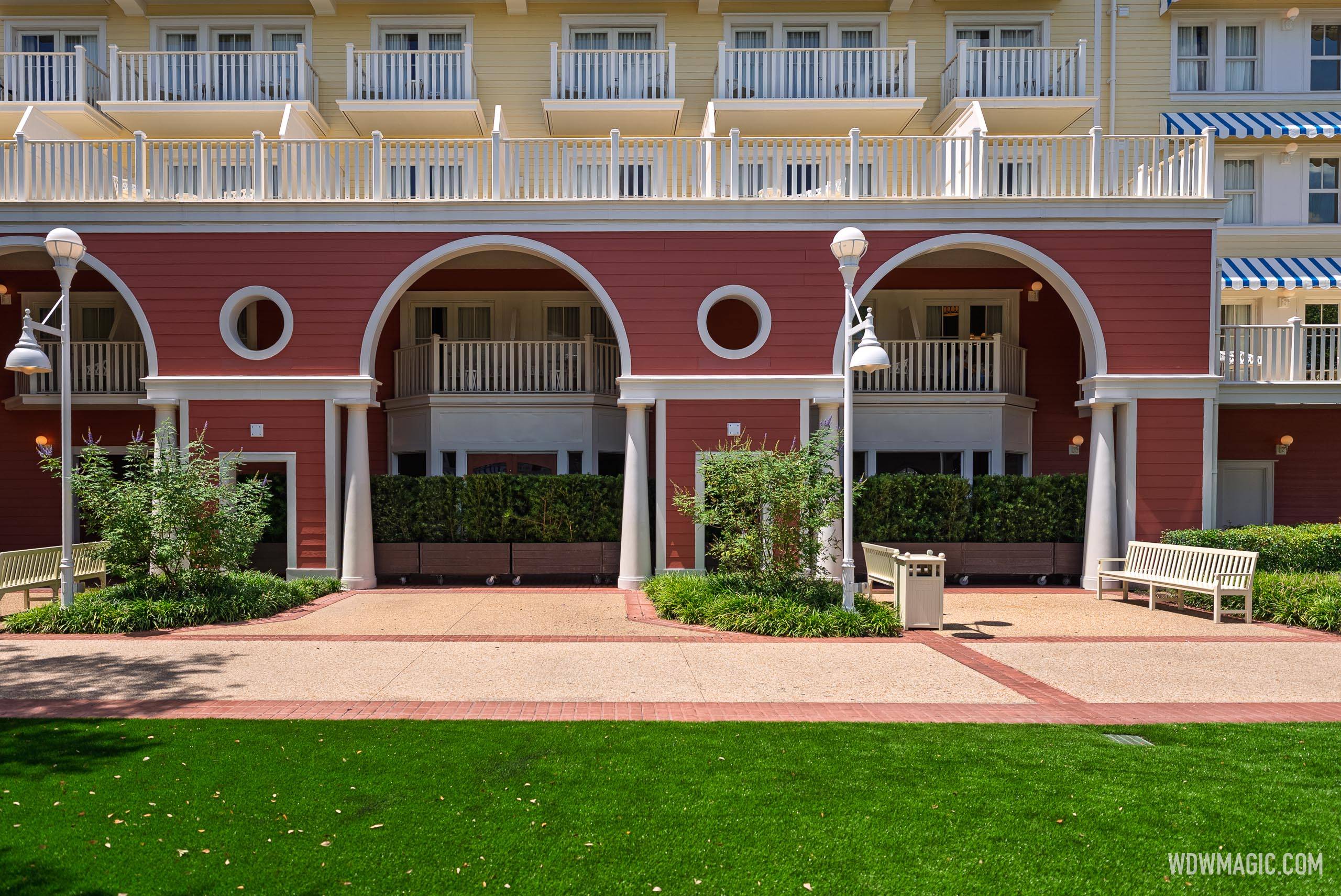 The red arcaded ground floor of a Disney's BoardWalk building with white columns, lamp posts and benches, the shop windows hidden behind rows of potted hedges