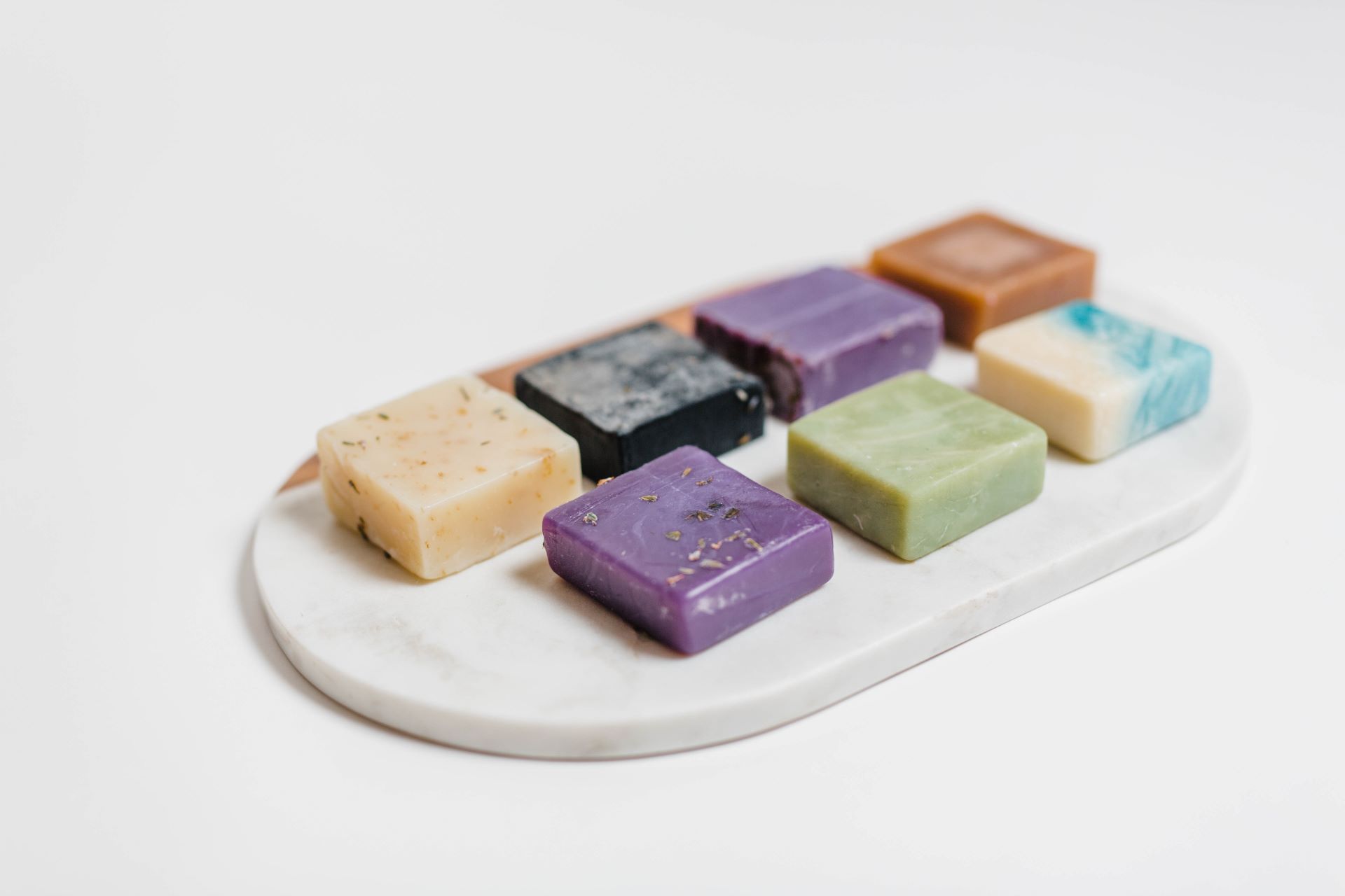 Seven square bars of handmade soap in cream, black, purple, green, brown and blue-and-white swirl on a white marble tray