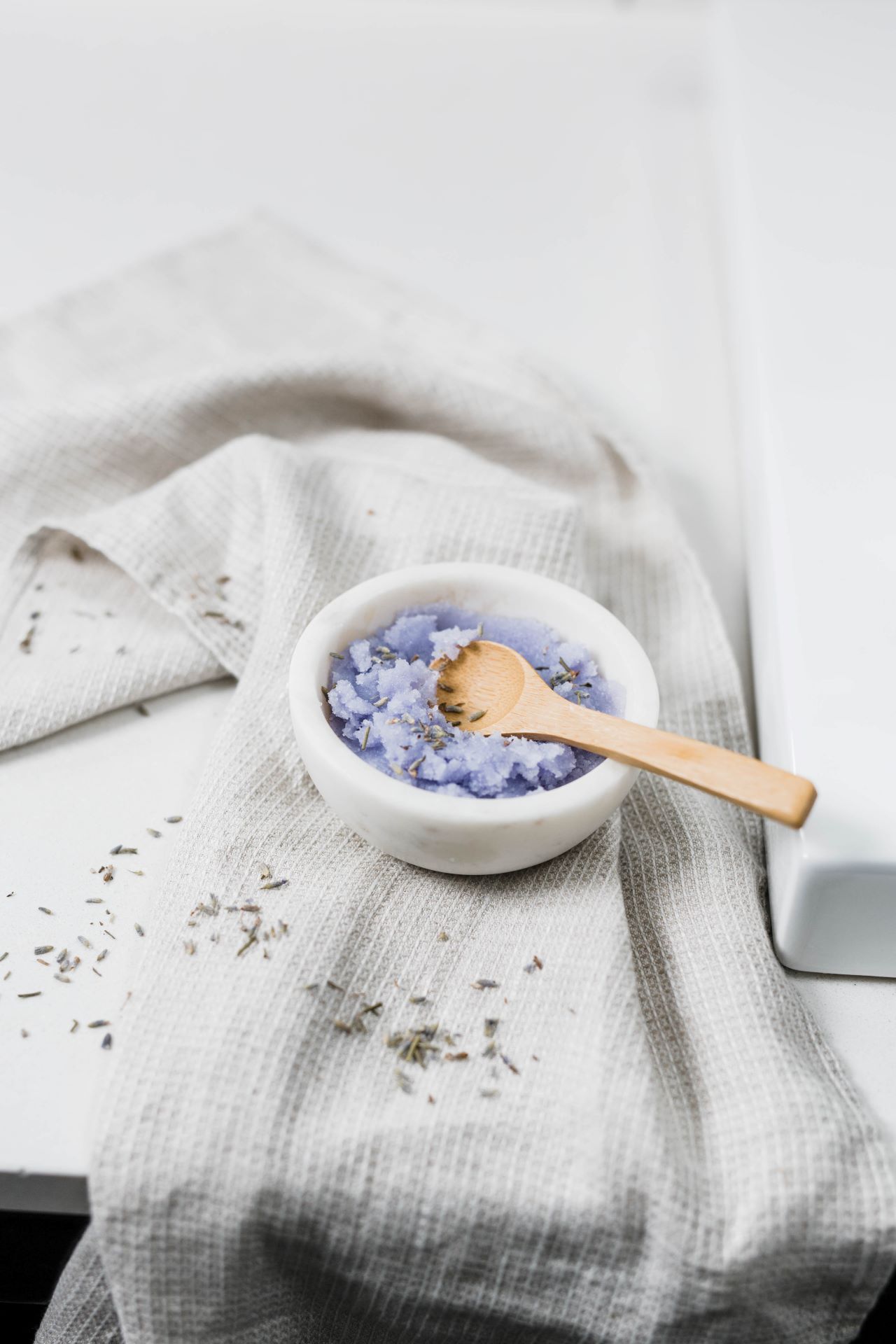A small white marble bowl of a lavender-colored bath product with a wooden spoon, set on a gray waffle-weave towel with dried lavender scattered around