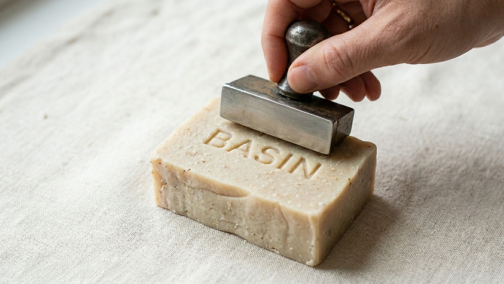 A hand presses a vintage metal stamp onto a cream-colored bar of soap embossed with the word BASIN, resting on linen