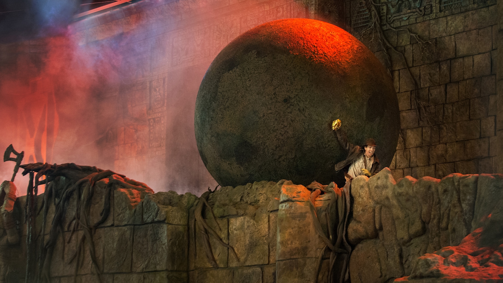 The opening scene of the Indiana Jones Epic Stunt Spectacular, with a huge round boulder looming behind Indiana Jones as he scrambles over a crumbling stone ledge holding up a golden idol, in front of a carved temple wall lit red and orange through smoke