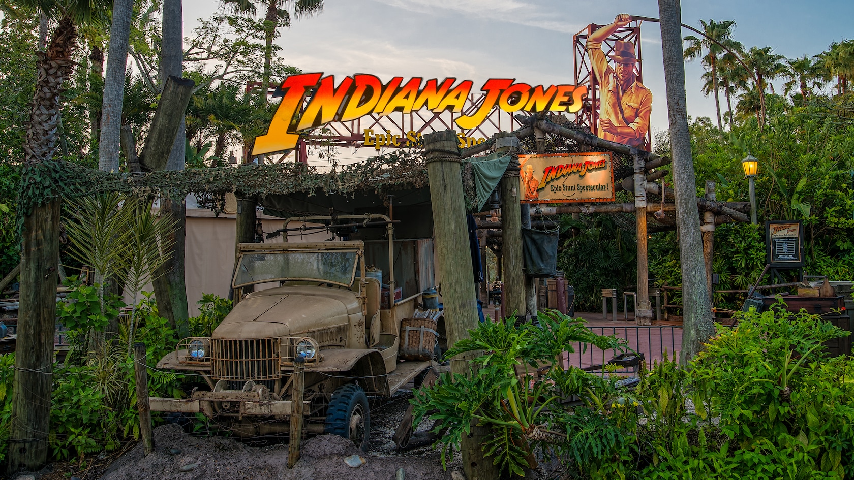 The Indiana Jones Epic Stunt Spectacular entrance at Disney's Hollywood Studios, with the orange and red INDIANA JONES marquee, a cutout Indy figure on a red steel tower, a weathered vintage truck under camouflage netting, a smaller show sign and tropical plants