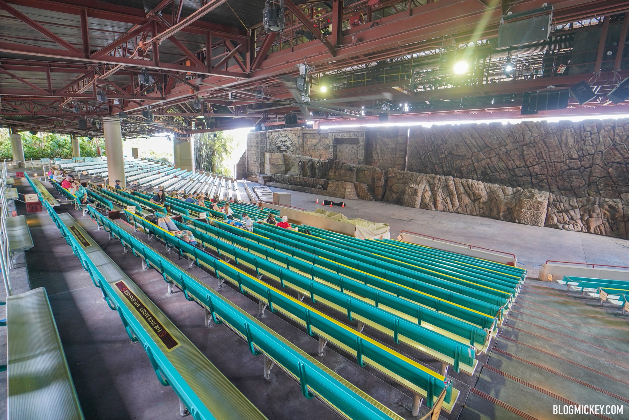 The Indiana Jones Epic Stunt Spectacular theater seen from the upper rows, with a scattering of guests resting on long teal bench seating under a covered roof held up by exposed steel trusses and lighting rigs, facing the stone-temple stage set