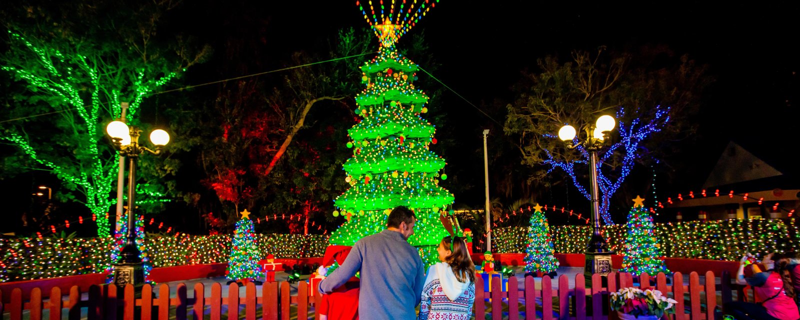 A man and a girl wearing reindeer antlers, with a child in a Santa hat beside them, stand at a red picket fence facing a tall tiered Christmas tree lit up in green at night, surrounded by smaller lit trees and trees wrapped in colored lights
