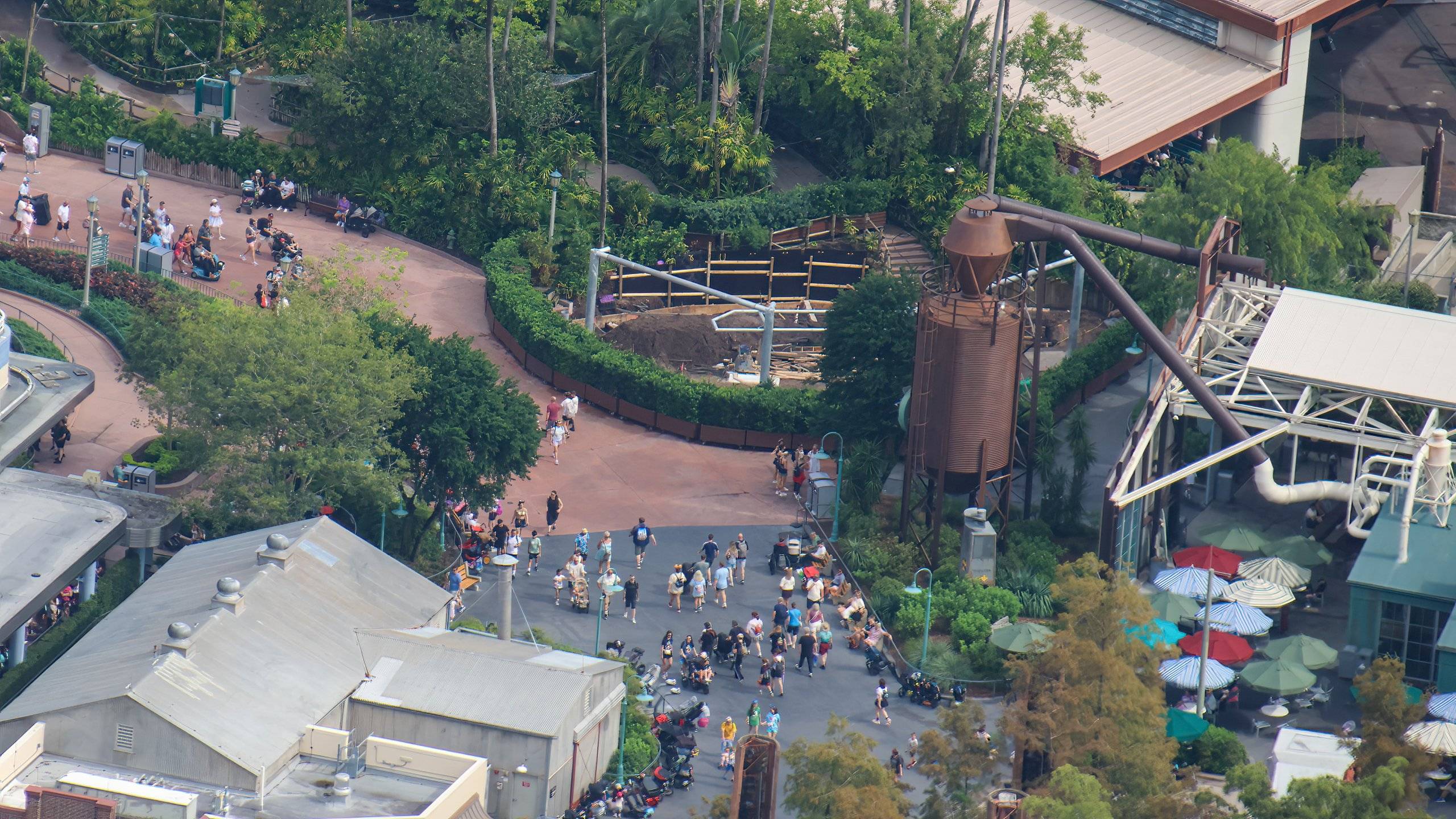 Aerial view of a walled construction site with a dirt floor, wooden form boards and metal posts, set behind a curving hedge beside a busy walkway, with a tall rusty silo-style tower and overhead ductwork to the right and a covered steel-framed structure at the edge of the frame
