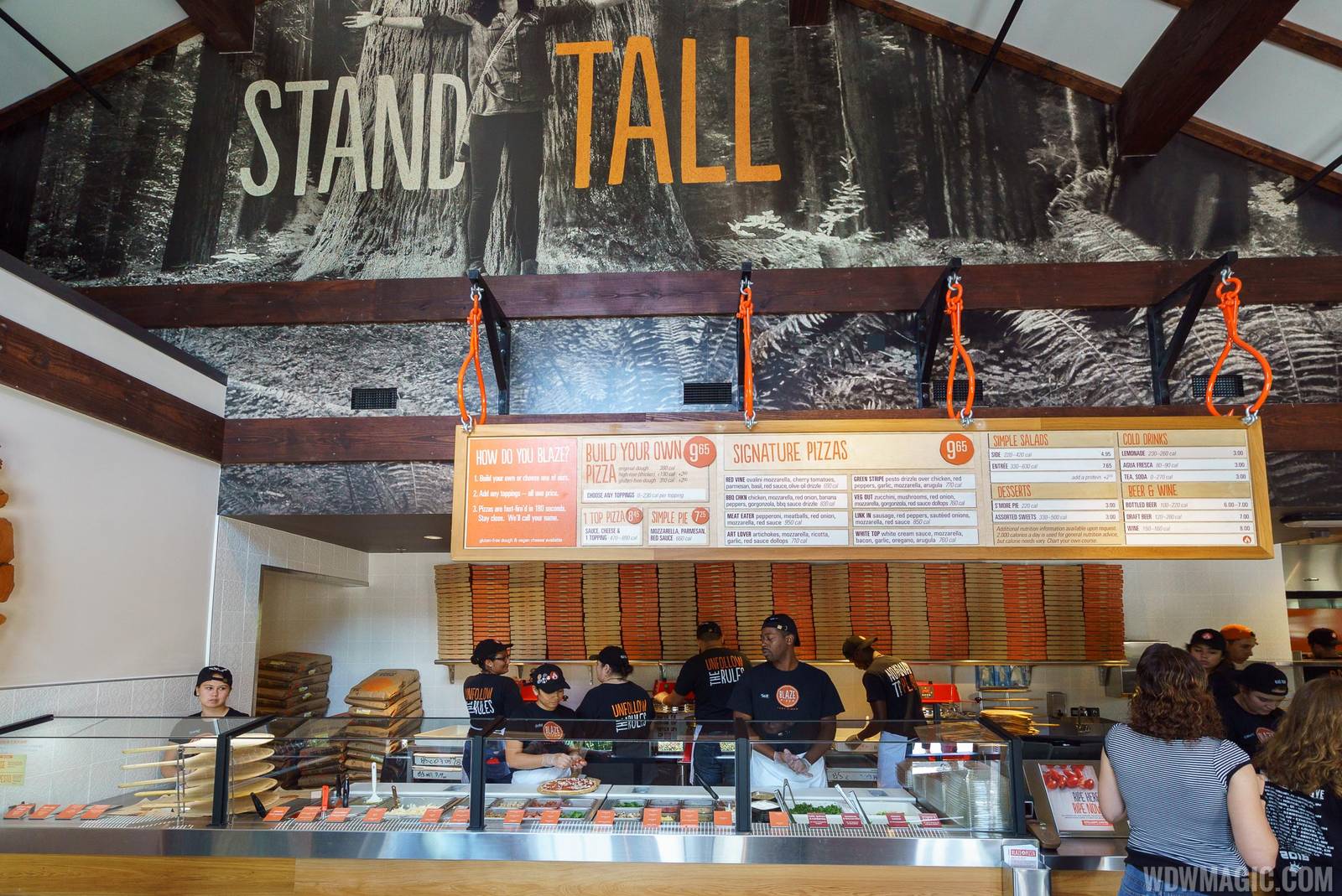 Staff in black Blaze caps working the pizza assembly line under a hanging menu board with Build Your Own Pizza, Signature Pizzas, Simple Salads and Desserts sections, below a large STAND TALL mural of a forest
