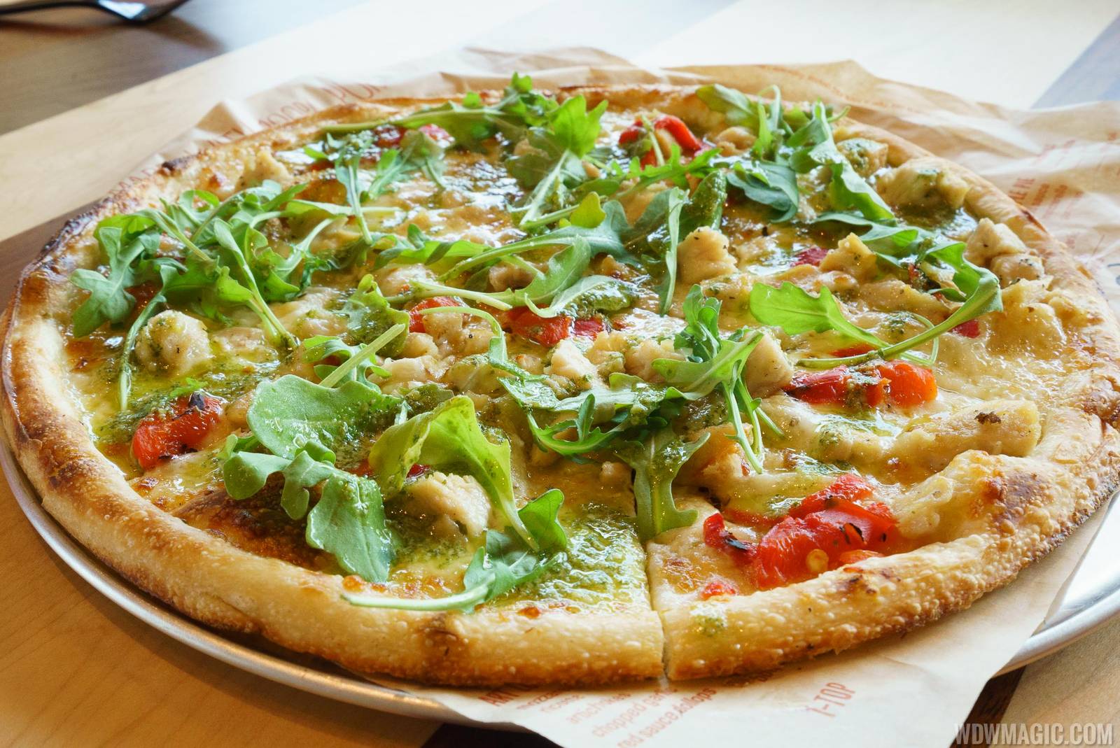 A whole thin-crust pizza on a metal tray and parchment, topped with chunks of chicken, roasted red peppers, melted mozzarella, a green pesto drizzle and scattered arugula