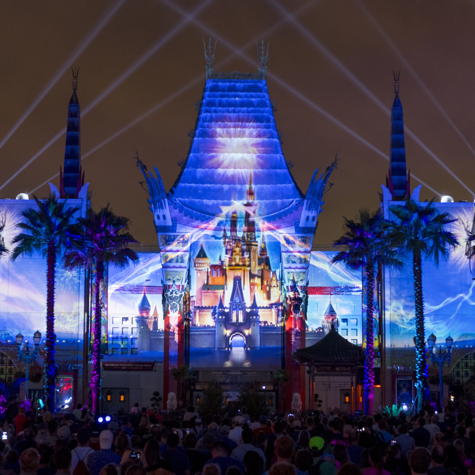 The Chinese Theater at Hollywood Studios at night with a castle projected across its facade, searchlight beams crossing the sky and a crowd watching from the street