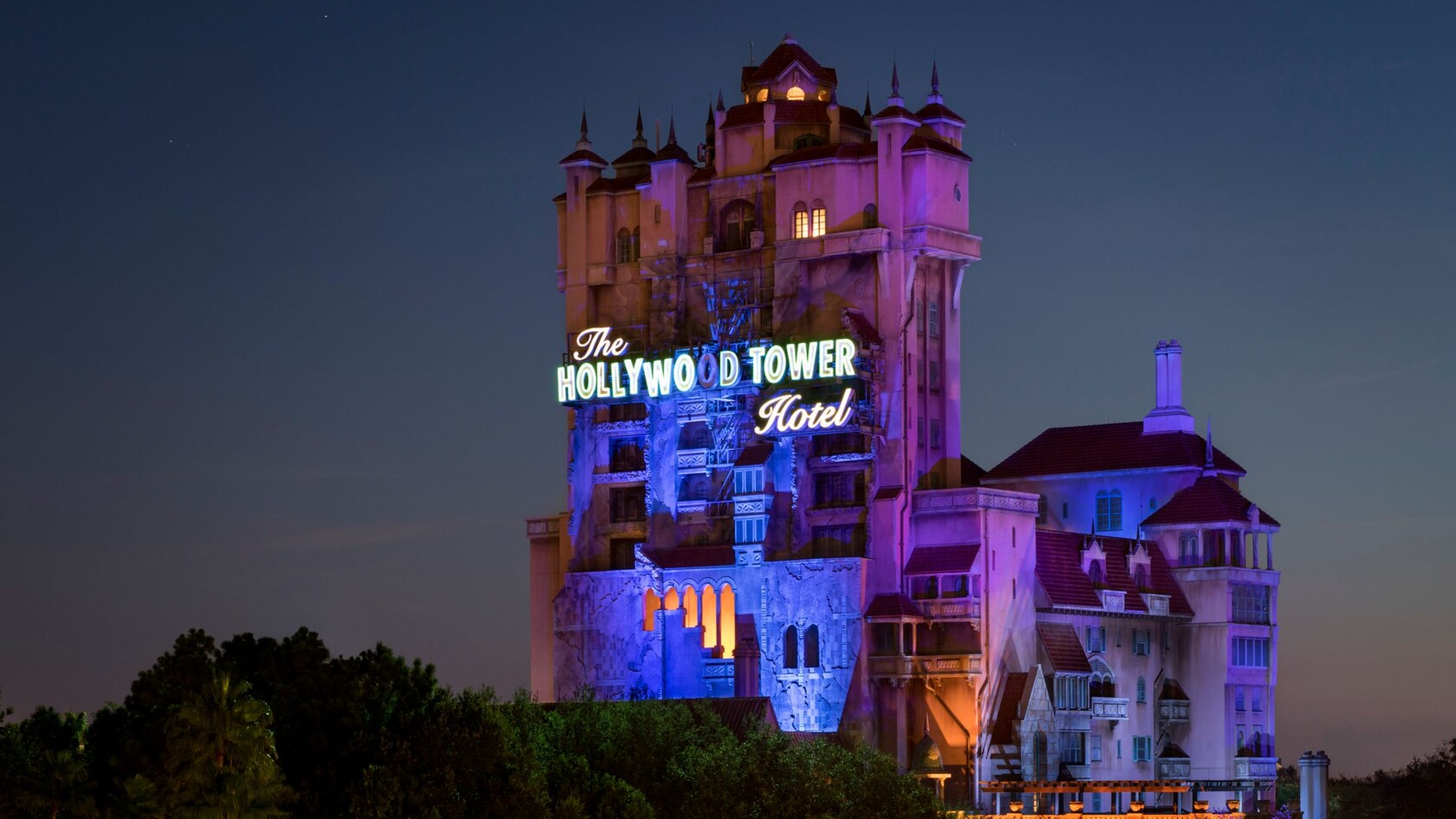 The Twilight Zone Tower of Terror at dusk, its Hollywood Tower Hotel sign lit up and the building washed in purple and blue light against a dark sky