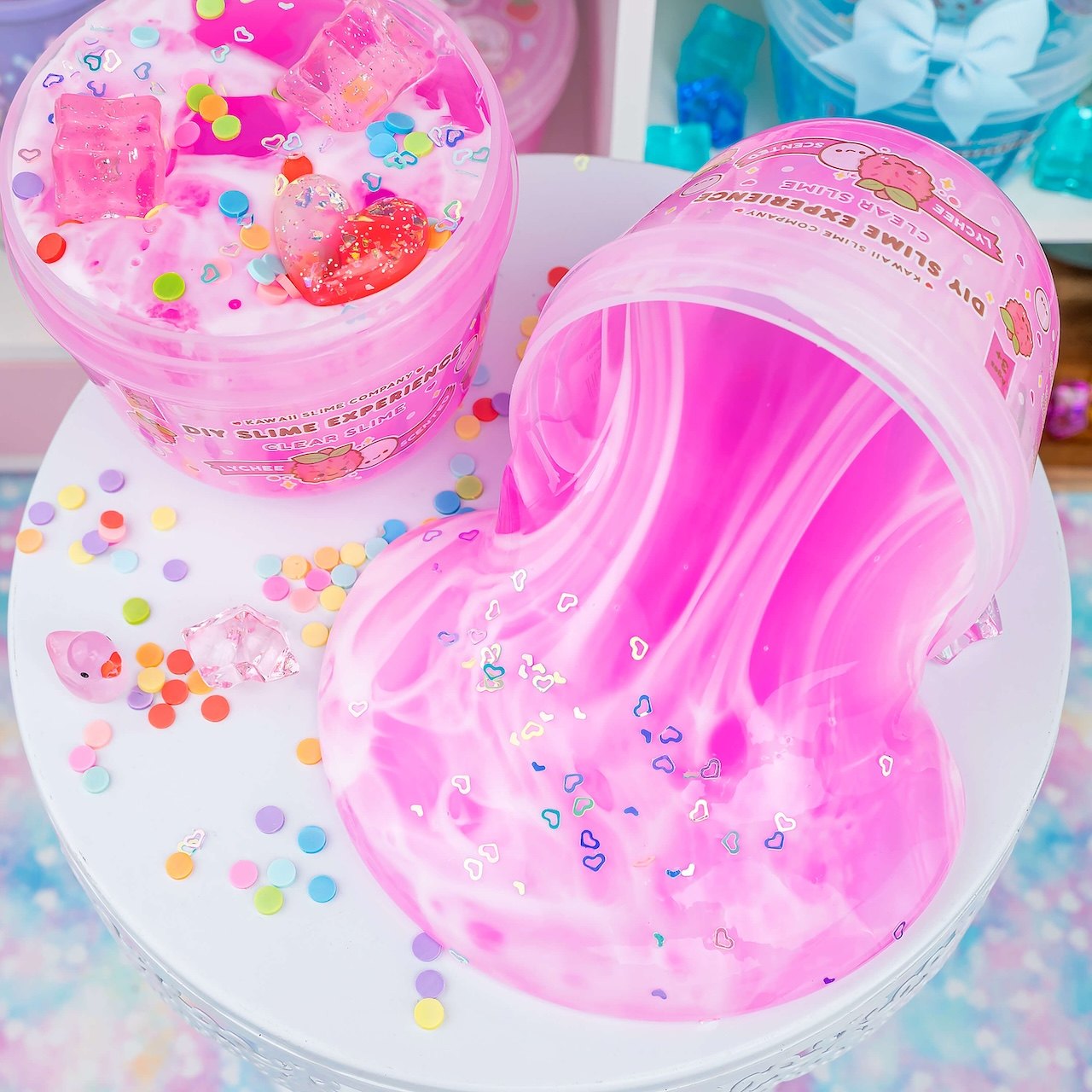 Two pink tubs of lychee-scented clear slime labeled DIY Slime Experience, one tipped over, surrounded by confetti and heart-shaped glitter
