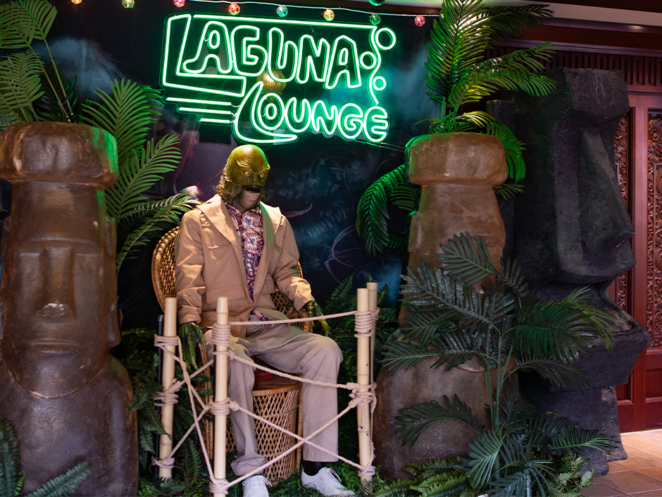 The Creature from the Black Lagoon in a tan suit and patterned shirt sits in a wicker chair behind a rope barrier, flanked by stone tiki heads and palms, under a green neon Laguna Lounge sign