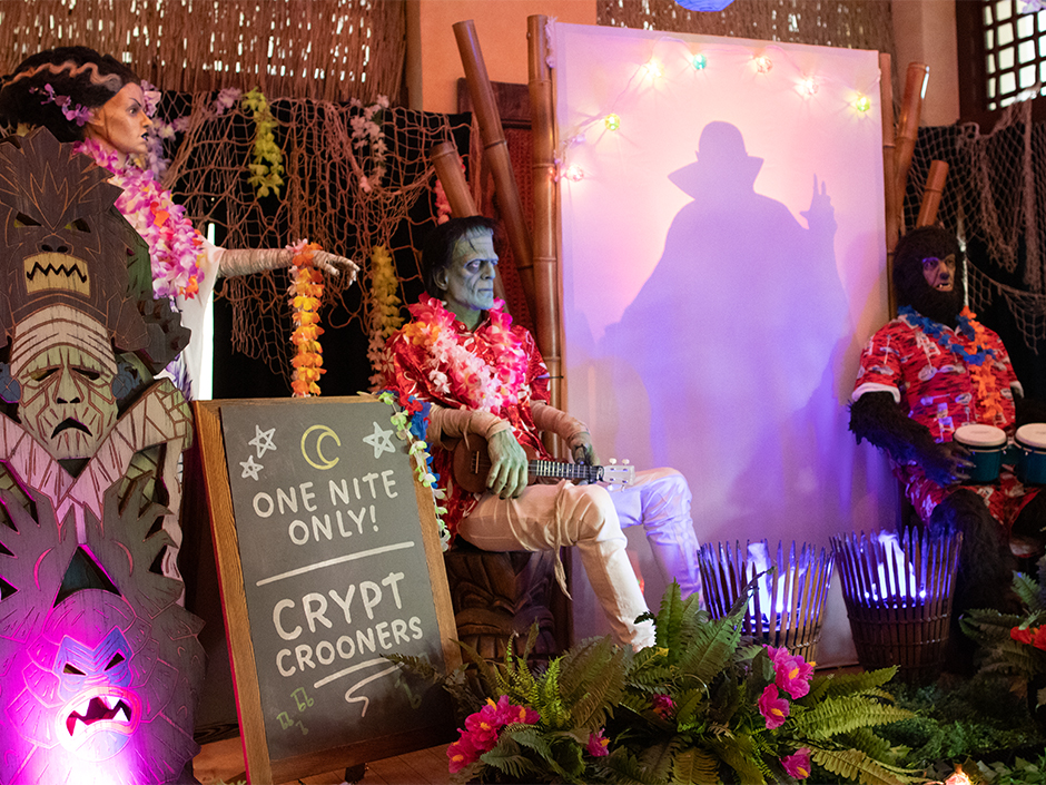Frankenstein's Monster with a ukulele, the Bride of Frankenstein in a white gown and the Wolf Man with bongos, all wearing leis, on a tiki stage beside a ONE NITE ONLY CRYPT CROONERS chalkboard and a backlit screen showing a shadowy silhouette