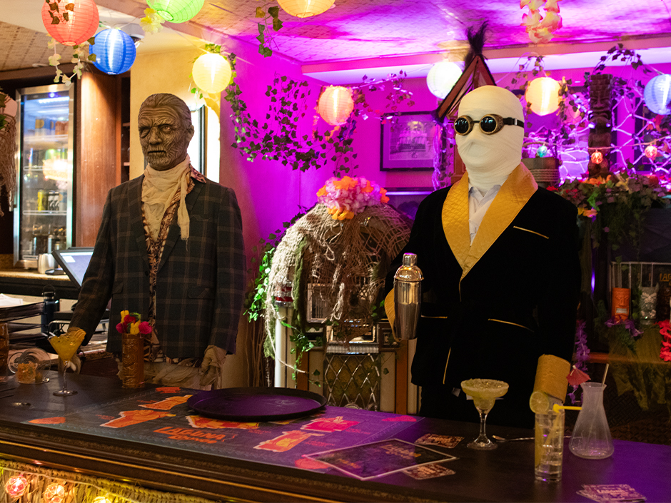 The Mummy in a plaid jacket and the Invisible Man in bandages, goggles and a black smoking jacket holding a cocktail shaker stand behind a bar under paper lanterns and purple light