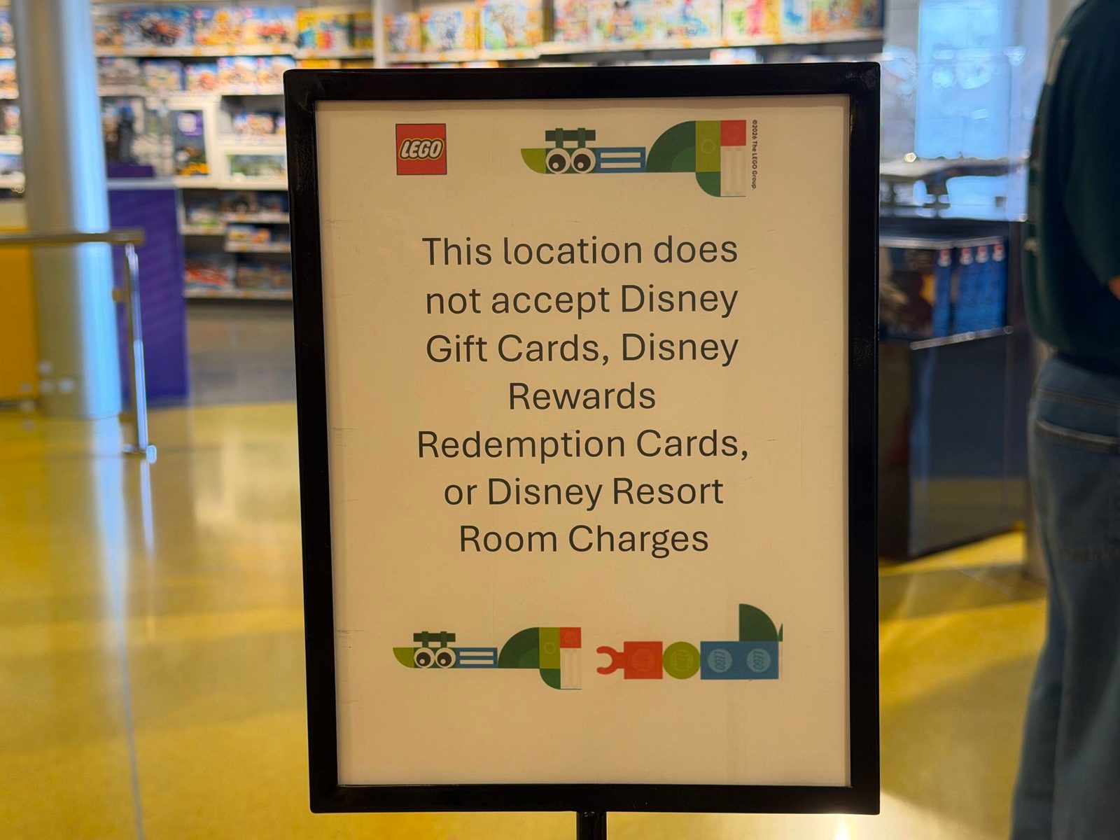 A framed sign on a stand inside The LEGO Store at Disney Springs, with a LEGO logo and brick graphics, reading: This location does not accept Disney Gift Cards, Disney Rewards Redemption Cards, or Disney Resort Room Charges. Shelves of LEGO sets fill the background.