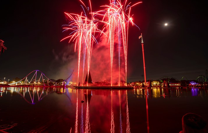 Red fireworks bursting over the lagoon at SeaWorld Orlando at night, reflected in the water, with a lit roller coaster track along the shoreline at left and a full moon in the sky