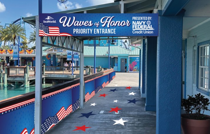 A covered walkway at SeaWorld Orlando under a blue banner reading SeaWorld Waves of Honor Priority Entrance, Presented by Navy Federal Credit Union, with American flag graphics along the railing and red, white and blue stars on the ground