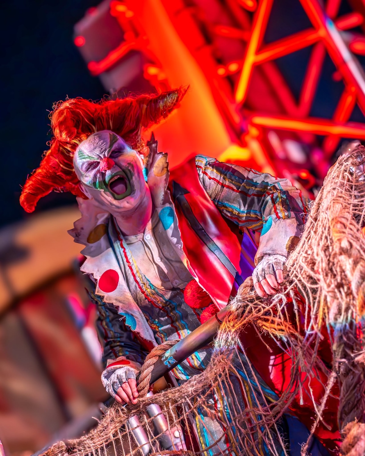 Jack the Clown, with wild orange hair and white face paint, screaming as he grips a rope net in front of glowing red neon scaffolding