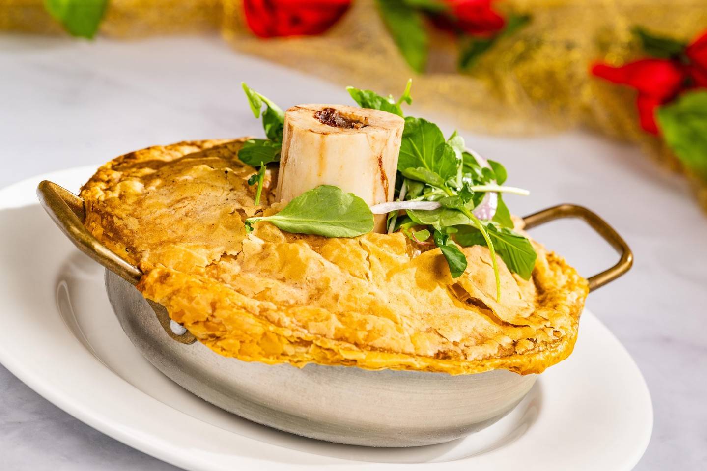 A golden pastry crust over a metal dish, topped with a roasted marrow bone and fresh greens, with red roses in the background
