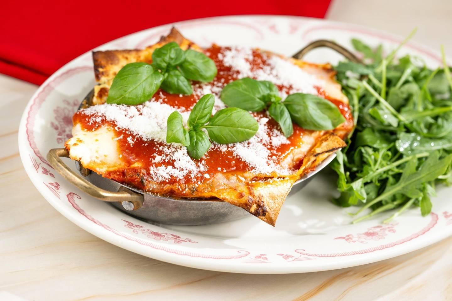 Lasagna alla Bolognese topped with tomato sauce, grated cheese and fresh basil in a small metal pan, with a side of arugula on a white plate with a pink rim
