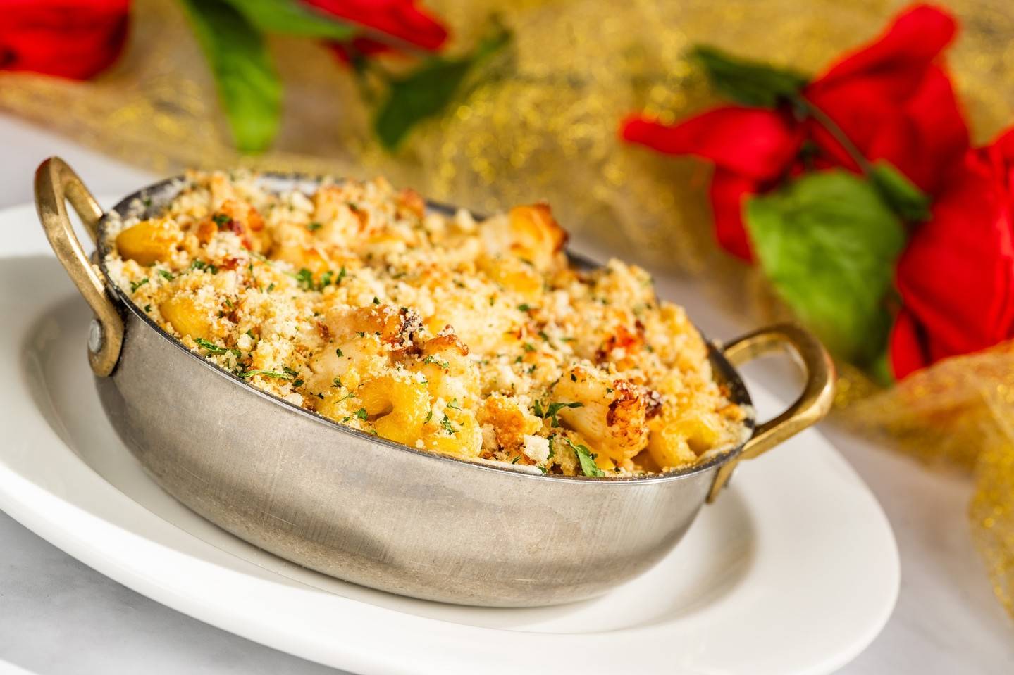 Macaroni with a golden breadcrumb crust in a two-handled metal dish on a white plate, with red roses behind it
