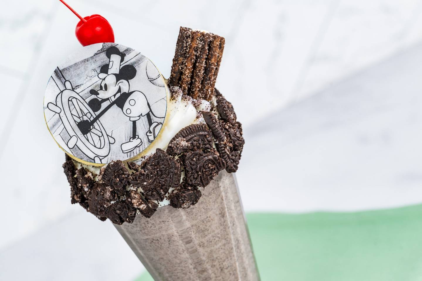 A cookies and cream milk shake topped with whipped cream, a cherry, a churro and a round sugar cookie printed with black and white Steamboat Willie Mickey at a ship wheel