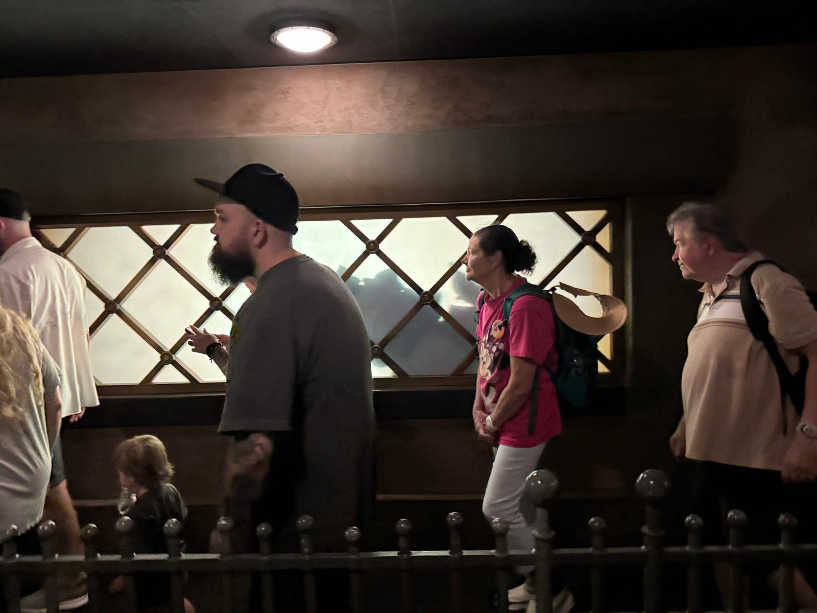 Guests walking through a dim, wood-paneled queue past a long illuminated window with frosted panes divided by a dark diamond lattice, with a faint shadow visible behind the glass