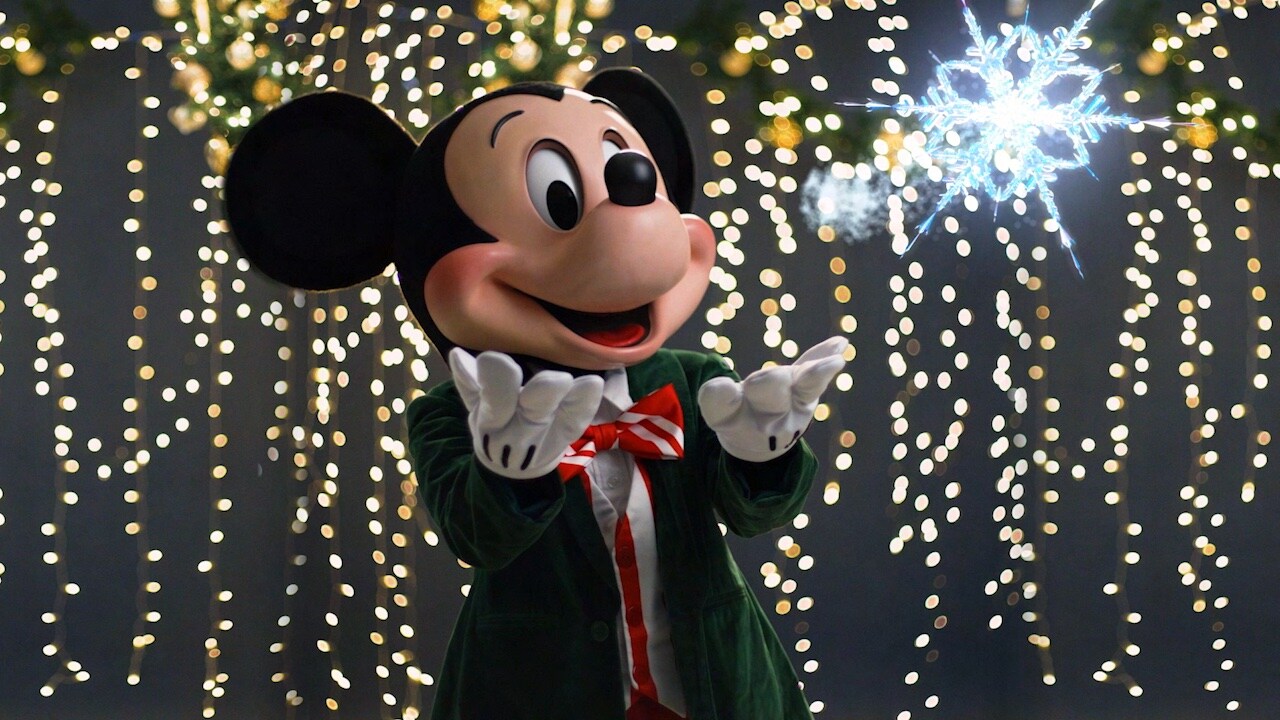 Mickey Mouse in a green velvet jacket and red striped bow tie, hands held out, in front of a curtain of warm string lights with a glowing snowflake at upper right