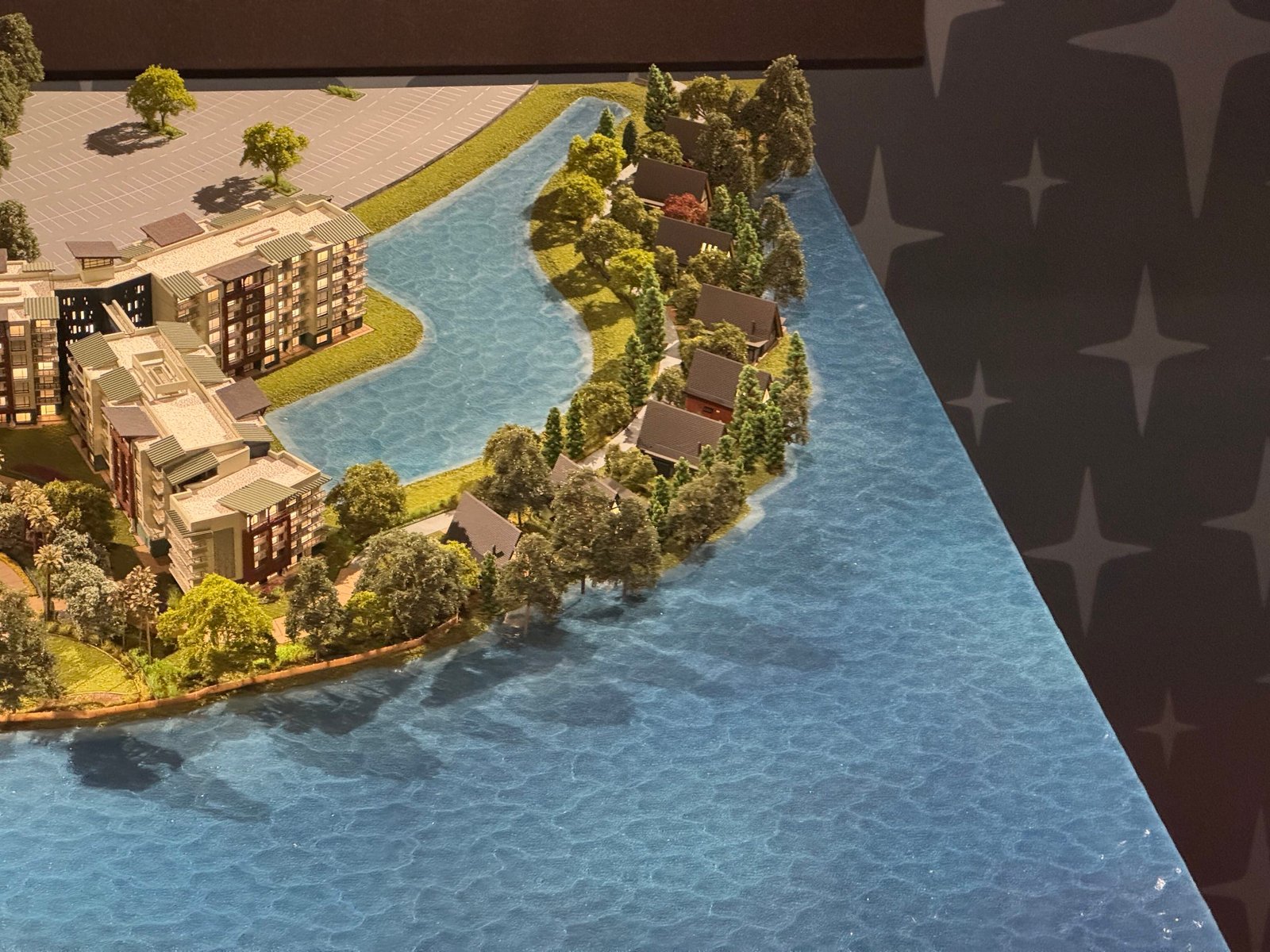 A corner of the Lakeshore Lodge model showing small dark A-frame houses among trees on a curving strip of land between two stretches of blue model water, with a hotel wing at left