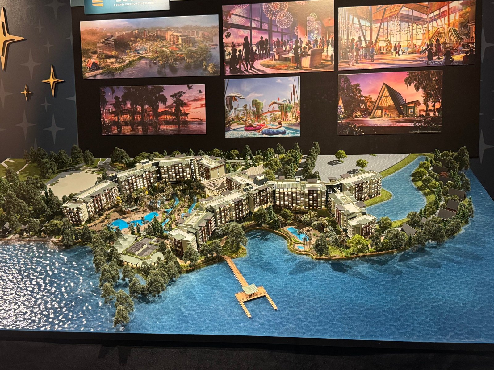 A large tabletop model of Disney's Lakeshore Lodge, with pale multi-story hotel wings, dense miniature trees and a pier reaching into blue model water, under a wall of six framed concept art pieces in Walt Disney Presents