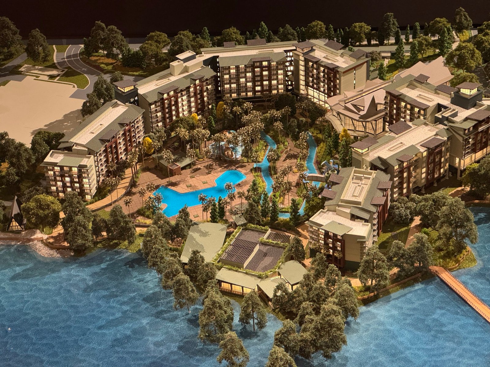 Close view of the Lakeshore Lodge model's main courtyard, with a bright blue free-form pool among palm trees and a lazy river curving past, surrounded by multi-story hotel wings