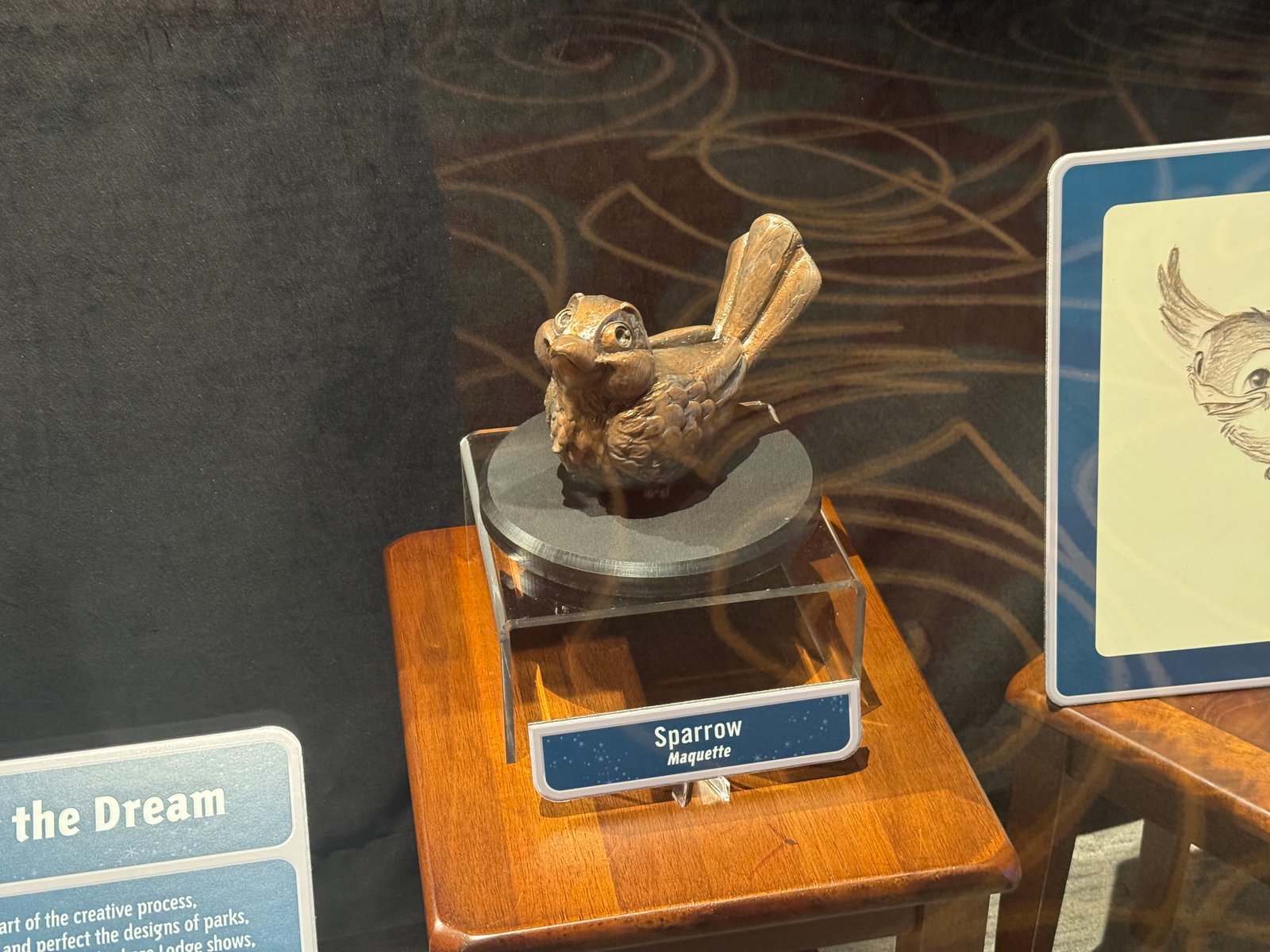 A small bronze-colored sparrow sculpture with large eyes and an upright tail on a round black base, on a wooden stand with a blue label reading Sparrow Maquette
