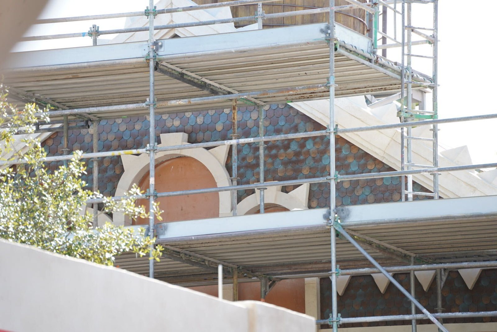 Behind scaffolding, a gable covered in small rounded shingles in blue, orange-brown and gray tones, above an arched window framed by a cream surround with pointed edges and a row of cream tooth-shaped trim below