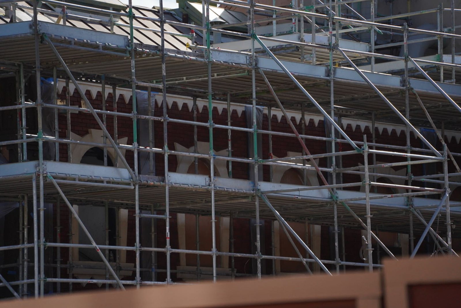 Scaffolding in front of a dark red brick building whose upper edge is lined with cream-colored trim cut into rows of pointed, tooth-like shapes, with arched window openings below